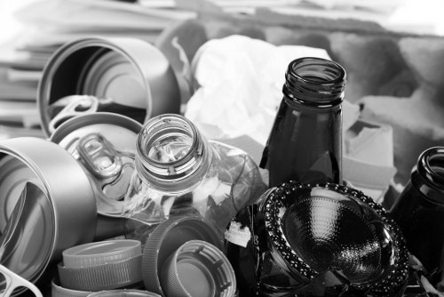 A collection of various waste items including several aluminum beverage cans with open tops, a clear plastic bottle with a cap, and a dark glass bottle, all situated among other miscellaneous rubbish such as plastic containers, a crumpled piece of paper or cloth, and a black object that appears to be a leather wallet or case, with a beaded chain or bracelet draped over it. These items are scattered on a surface, potentially a driveway or pavement, in an outdoor setting. The background shows more indistinct rubbish or packaging materials, indicating a typical scene of waste awaiting collection or disposal. This image relates to rubbish removal services offered by Palmers Green House Clearance, situated in or near Palmers Green, North London, with a focus on clearing household waste, including cans, bottles, and general clutter, in an urban or suburban environment.