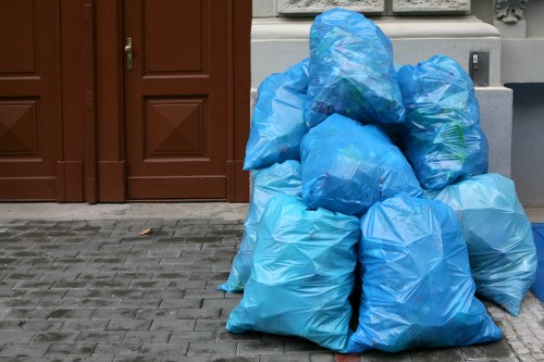 A collection of several large blue rubbish bags, made from a glossy plastic material, are piled on a paved pavement area in front of a brown wooden door and matching wooden cabinet doors. The bags appear to be filled with household waste or debris, with some slightly bulging or irregular in shape, indicating they are full. They are positioned near a grey wall and a metal utility box, suggesting a residential or commercial property in a suburban setting. The pavement consists of rectangular grey bricks, providing a stable surface for the waste collection. The scene captures the typical outdoor rubbish disposal scene, which Palmers Green House Clearance regularly manages on residential drives or front gardens as part of their house clearance and waste removal services. The lighting is natural, with no strong shadows or artificial illumination, emphasizing the practical and neutral nature of the cleanup process in the local area.