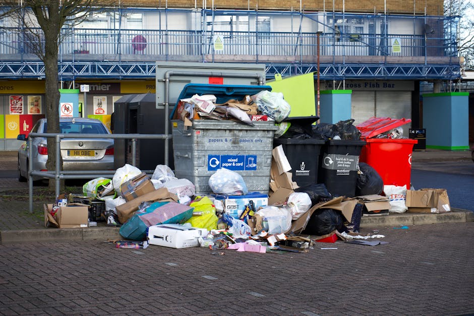 A middle-aged man with short dark hair, dressed in a dark T-shirt and matching trousers, is seen disposing of rubbish at an outdoor public waste receptacle. He is leaning forward, placing a white plas